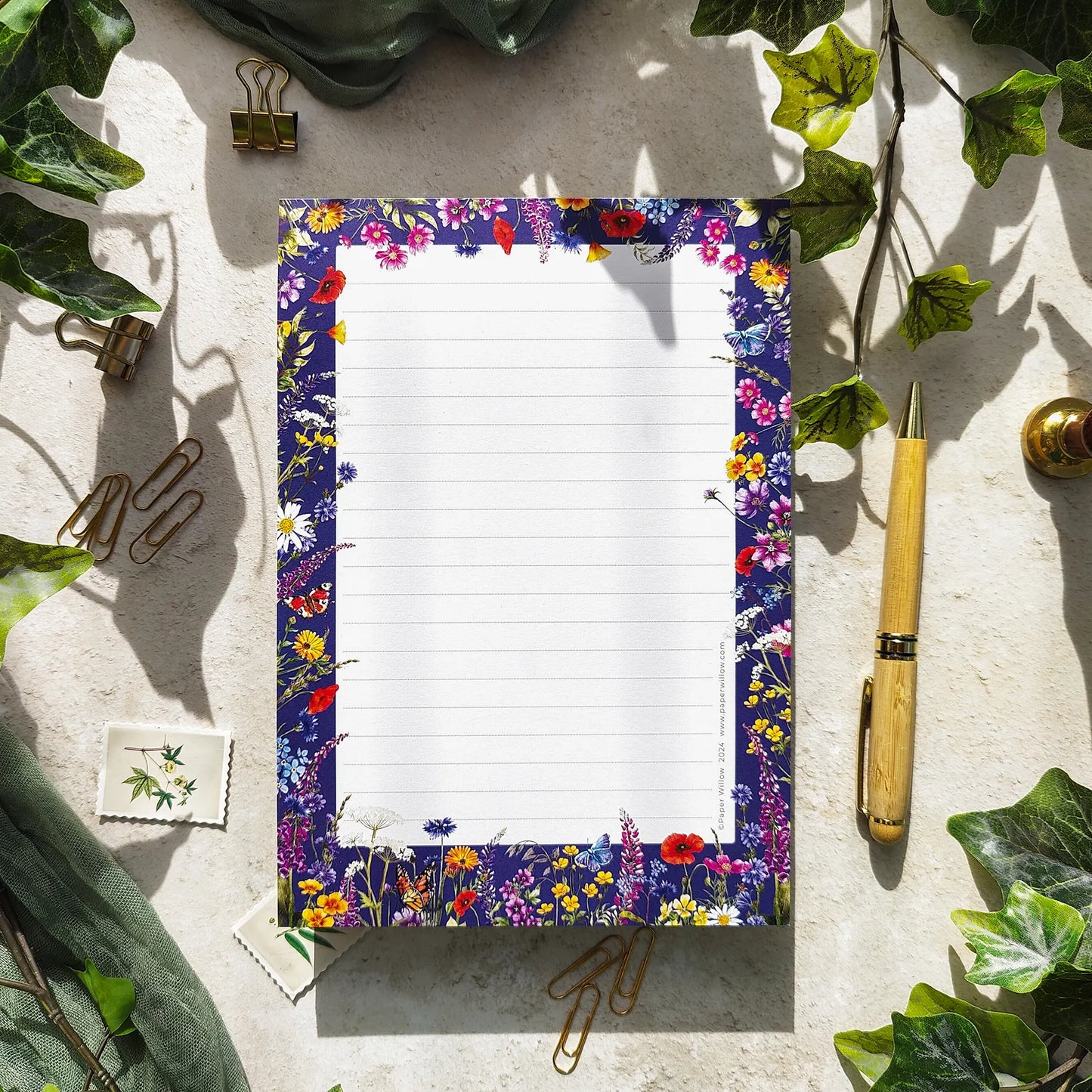 Floral border notepad on a textured surface with greenery and stationery items.