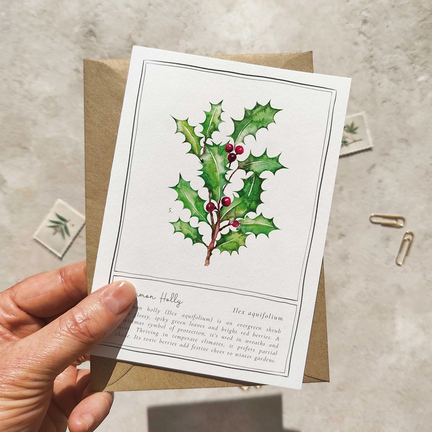 Hand holding a card with holly leaf design and text on a neutral background