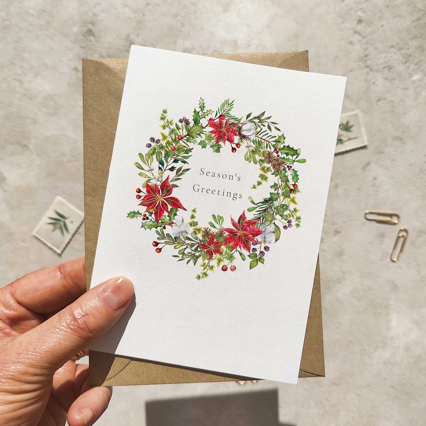 Hand holding a Christmas card with floral wreath design and 'Season's Greetings' text.