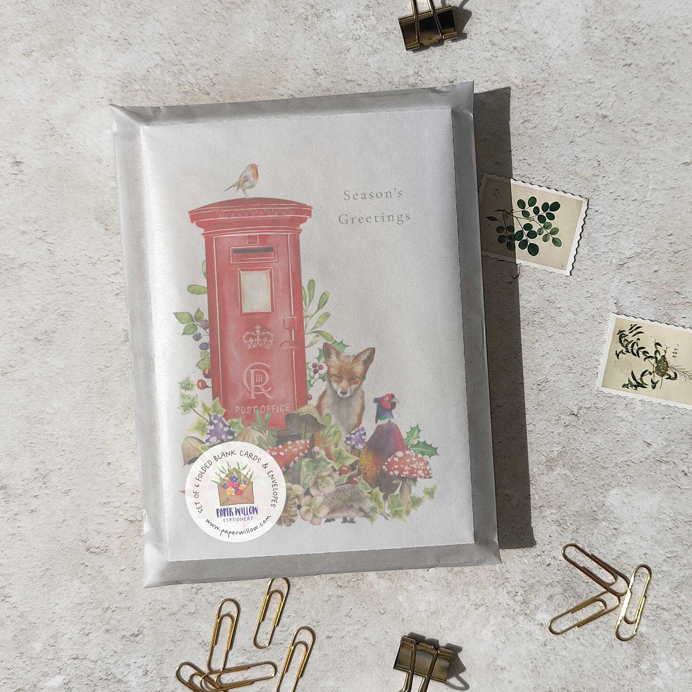 Greeting card with a red postbox and fox illustration on a textured surface with paper clips.