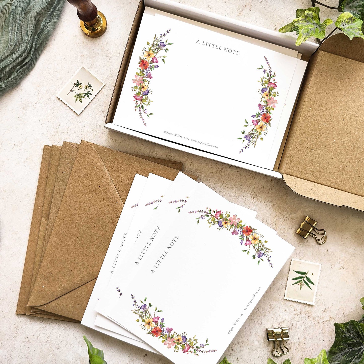 A set of blank notelets with floral designs and plain kraft envelopes, displayed in an open box with a leafy decoration.