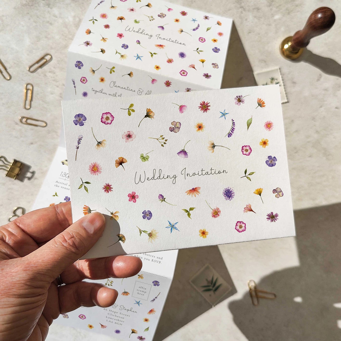 Floral wedding invitation card held by a hand with stationery items on a light surface.
