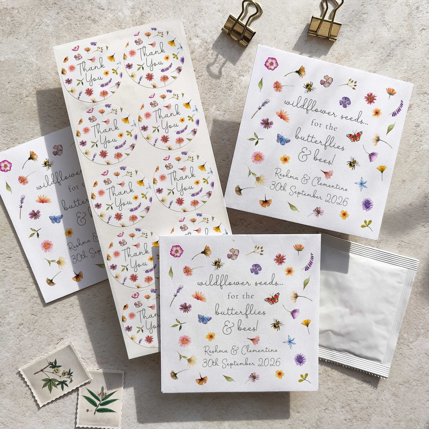 A collection of pressed wildflowers seed packets with floral designs, including personalized details such as names and dates. The packets are displayed on a textured surface with a butterfly and bee motif.