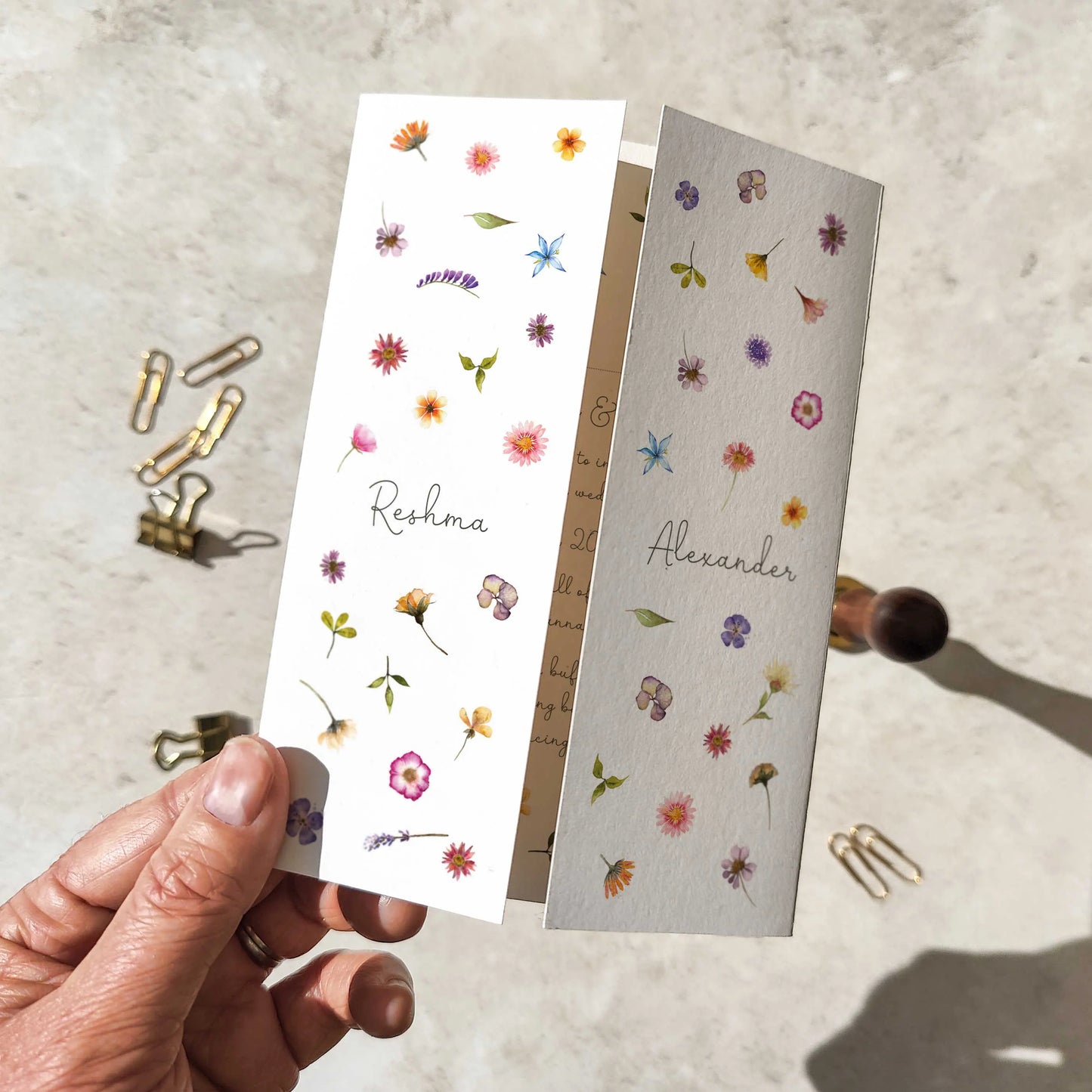 Wildflower wedding invitation held by a hand on a textured surface with paper clips.