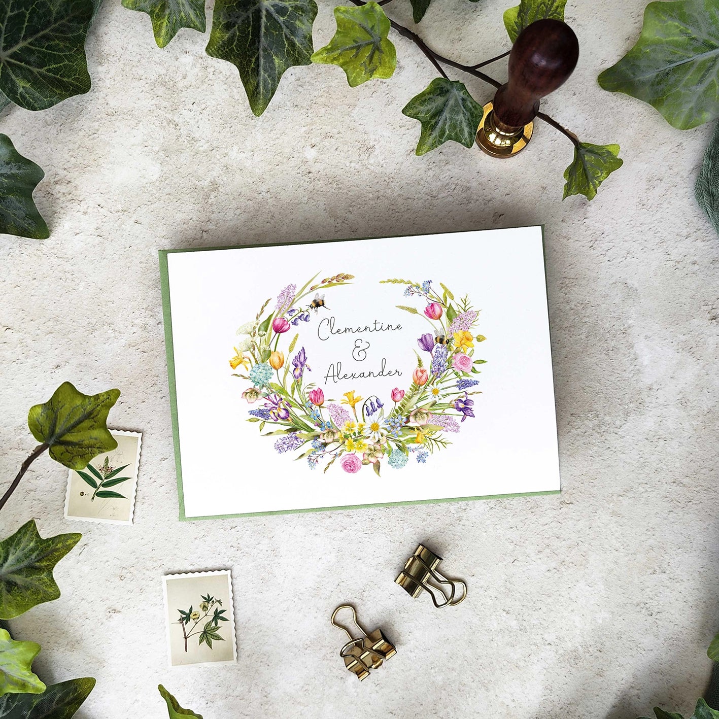 Floral wreath wedding invitation card with names on a textured surface with leaves and stationery items.