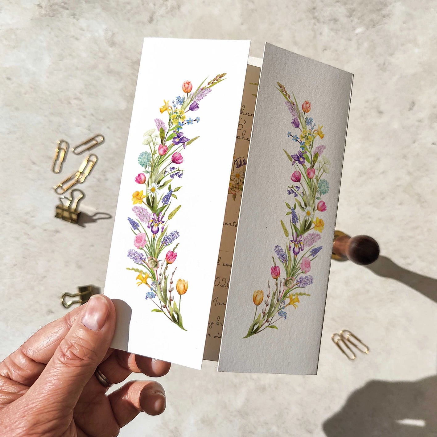 Spring Floral wedding invitation held by a hand with paper clips on a textured surface