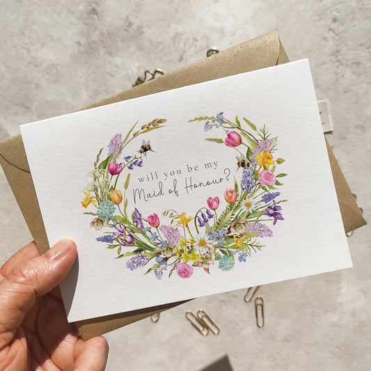 Floral wreath card asking 'Will you be my Maid of Honour?' held by a hand with a beige envelope.