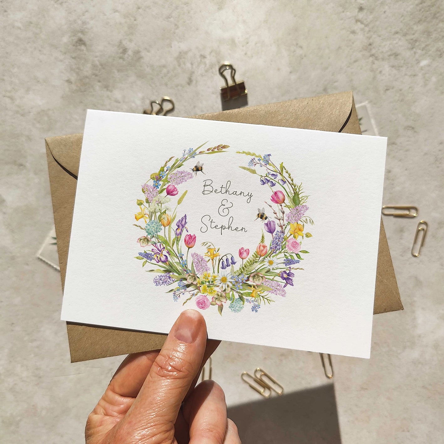 Folded wedding invitation with spring floral design and 'Bethany & Stephen' text, held over a textured surface.