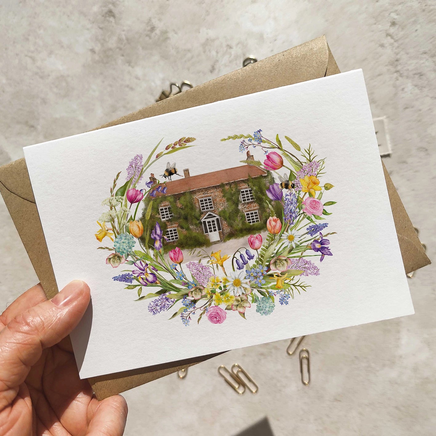 Wedding invitation with spring floral design and house illustration held by a hand on a neutral background