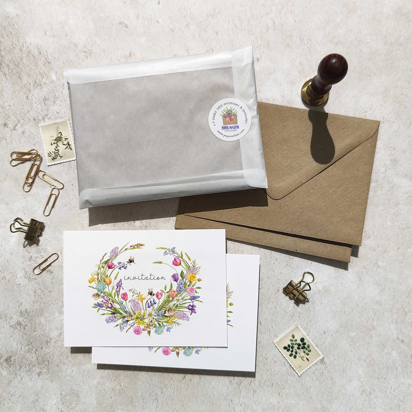 Invitation card with floral design, gray envelope, brown envelope, and stationery items on a textured surface.