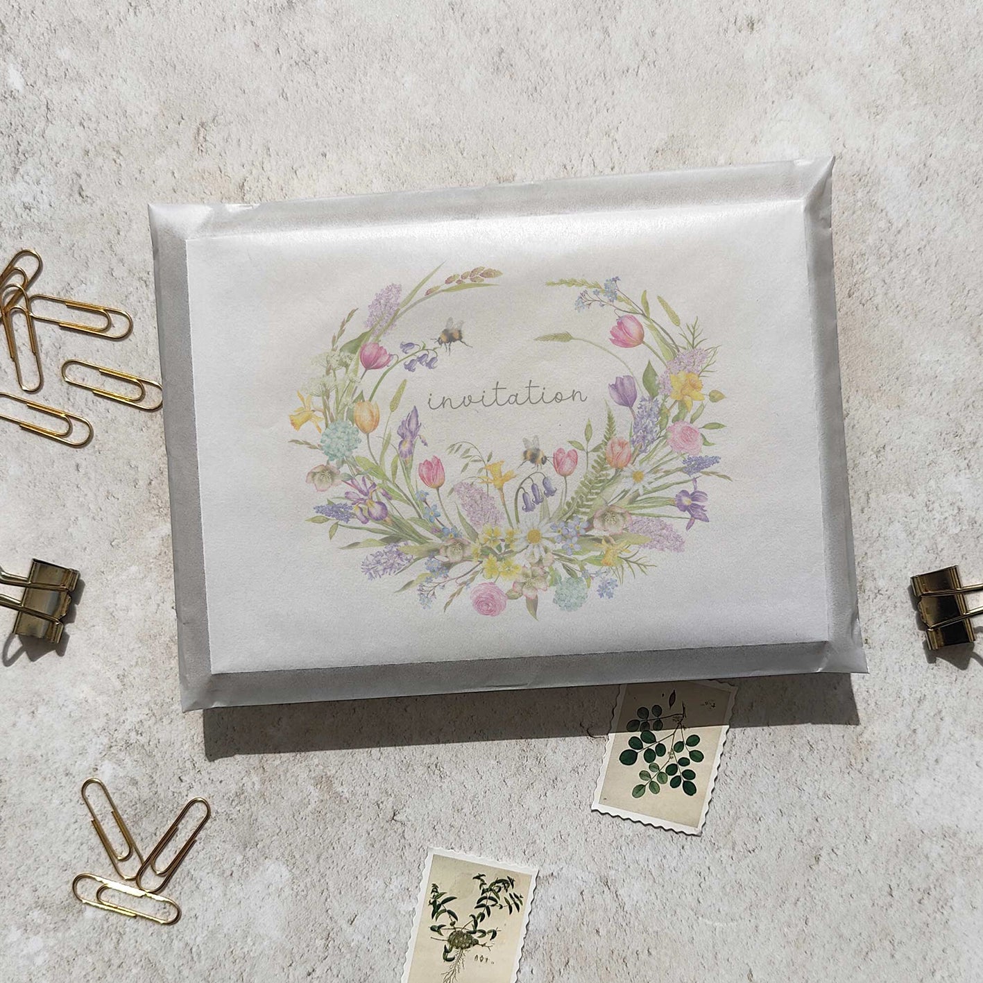 Invitation card with spring floral wreath design on a textured surface with paper clips and a small card.
