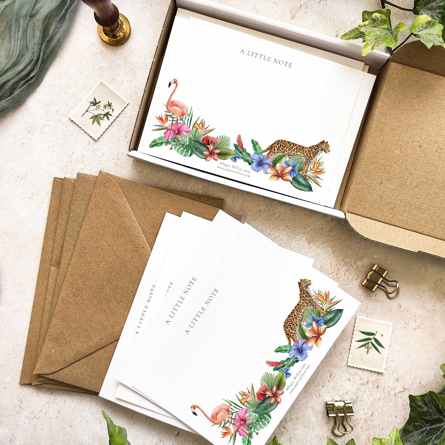 A set of blank kraft envelopes with a tropical watercolor floral design, displayed in an open box with a few envelopes visible outside. The background includes a small note card with a floral design and a keychain.