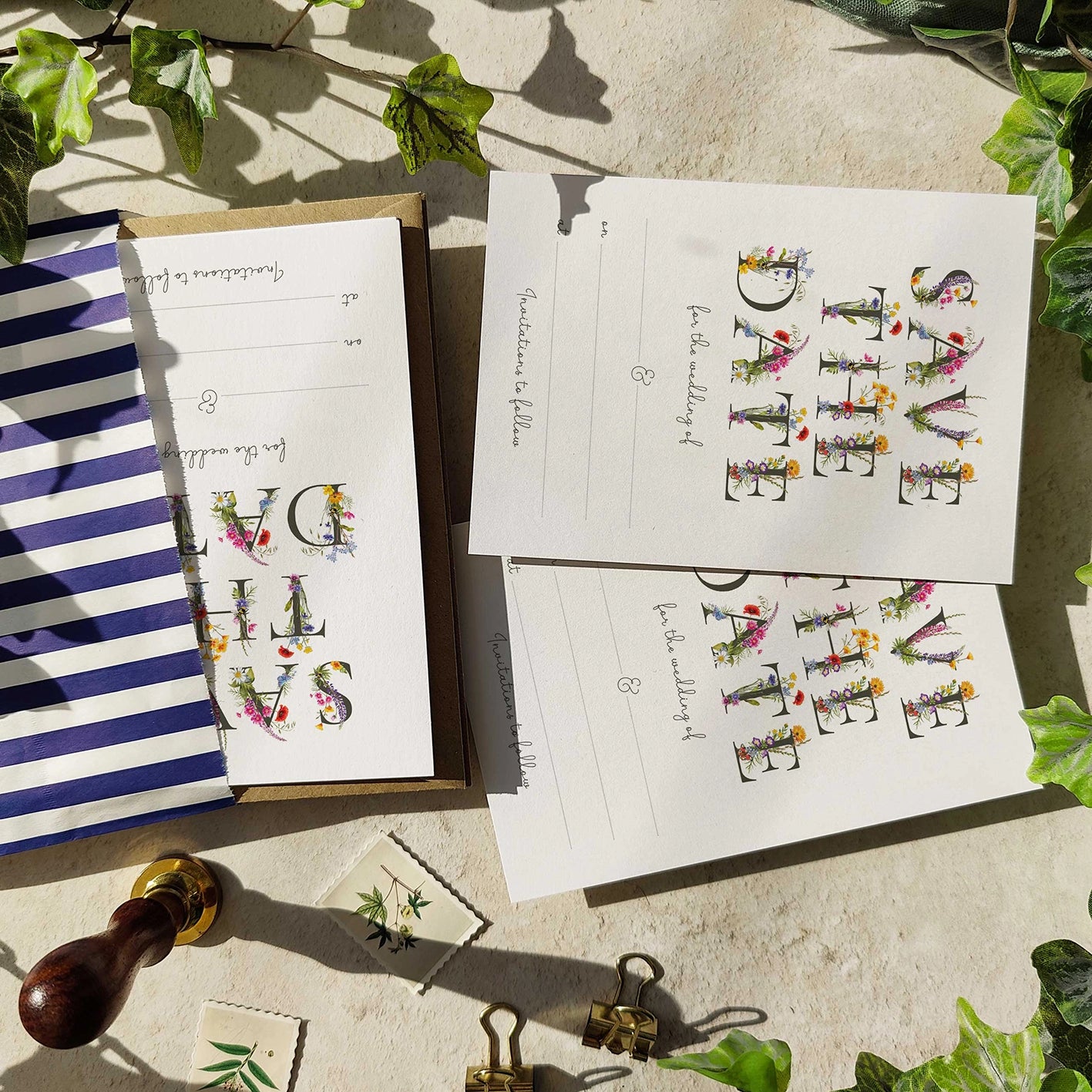 Stationery items with floral letter designs on a textured surface with greenery.