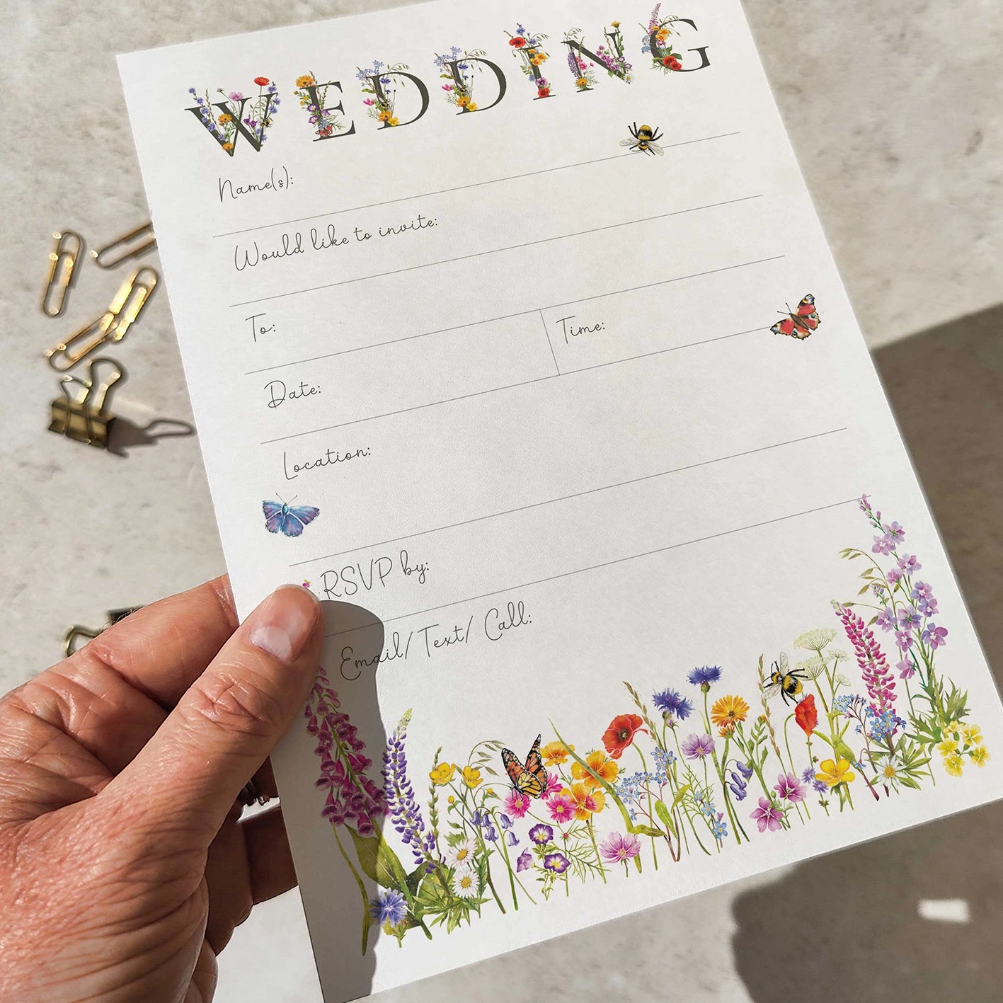 Hand holding a wedding invitation card with floral designs and stationery items on a concrete surface.