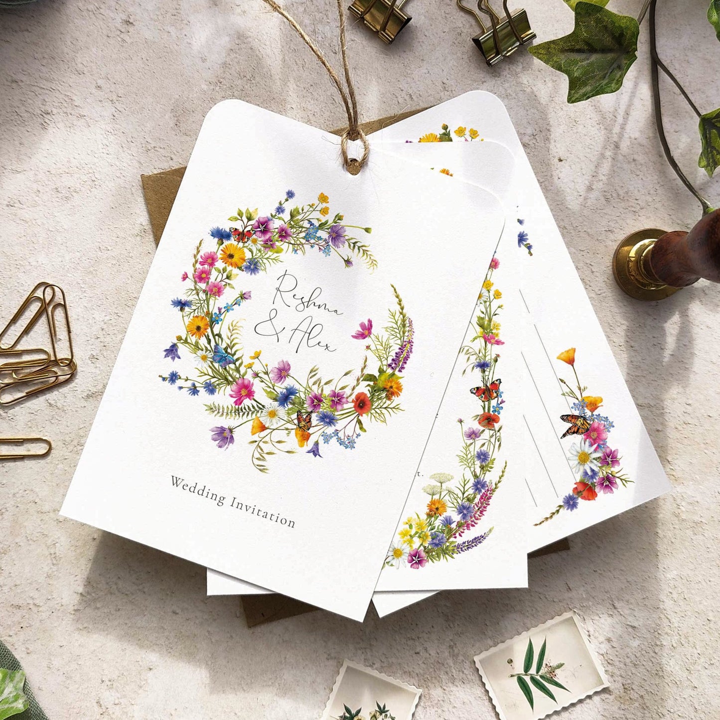 A set of hand-tied wedding invitations with floral designs including cornflowers, poppies, and buttercups, displayed on a table with a RSVP postcard, a pen, and some decorative leaves.