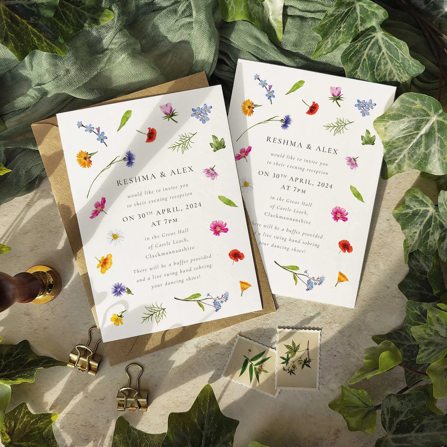 Wedding invitations with floral designs on a stone surface surrounded by green leaves.