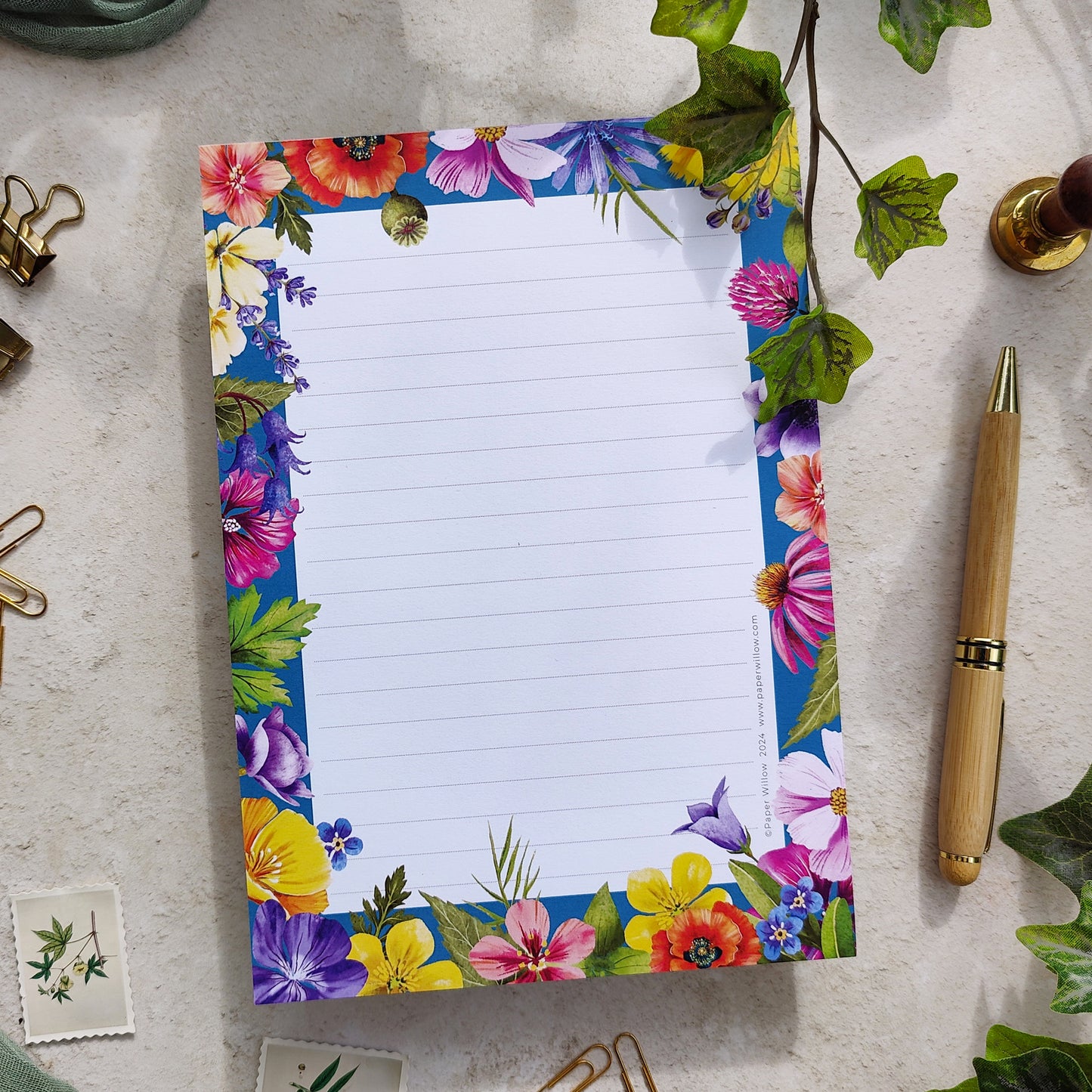 A colorful A5 notepad with a floral design on the cover, displayed on a table with a pen and some decorative items in the background.