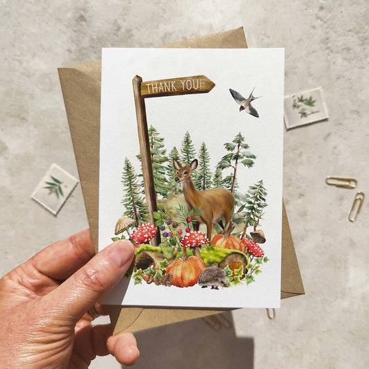 Hand holding a greeting card with nature-themed design featuring a deer, trees, and pumpkins.