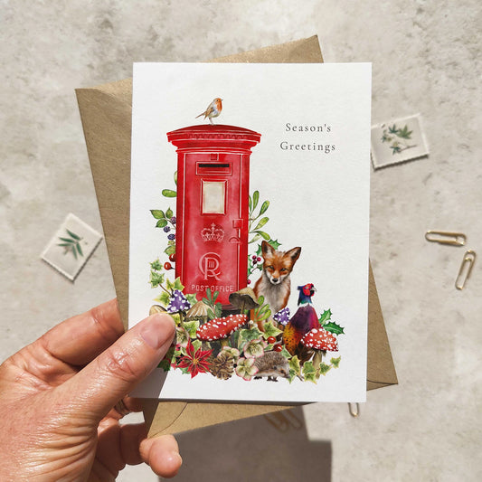 Hand holding a Christmas card with a red postbox, fox, and nature elements on a textured surface.