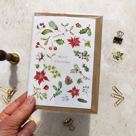 Hand holding a Christmas-themed greeting card with nature illustrations on a light surface.