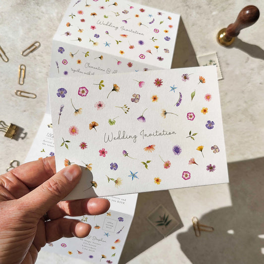 Floral wedding invitation card held by a hand with stationery items on a light surface.