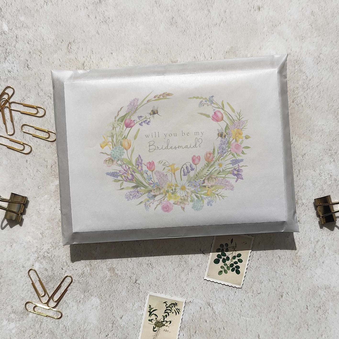 Floral wreath card asking 'will you be my bridesmaid?' on a textured surface with paper clips.