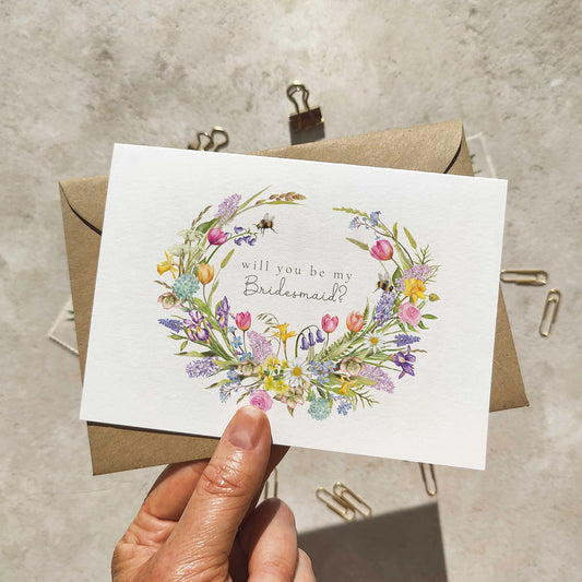 Floral card asking 'Will you be my Bridesmaid?' held over a concrete surface.