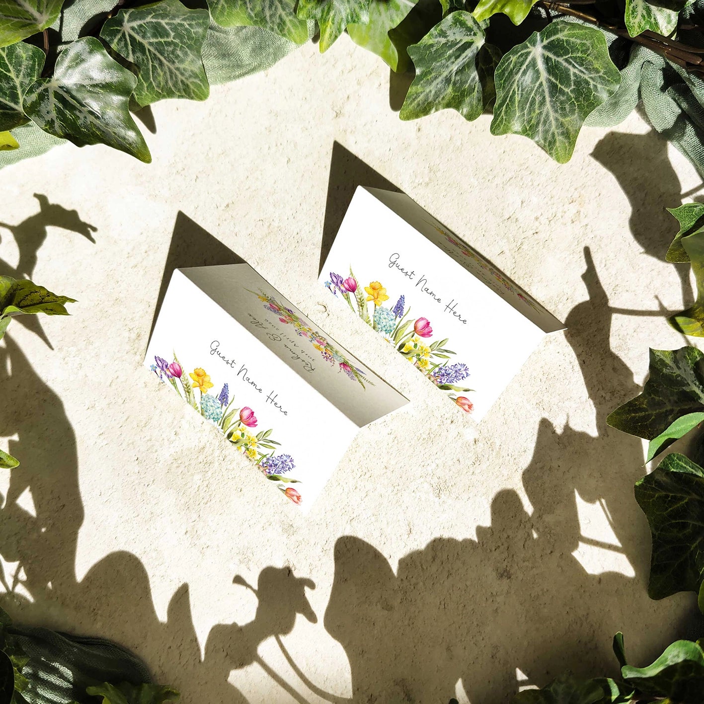 Two place cards with spring floral designs on a textured surface surrounded by green leaves.