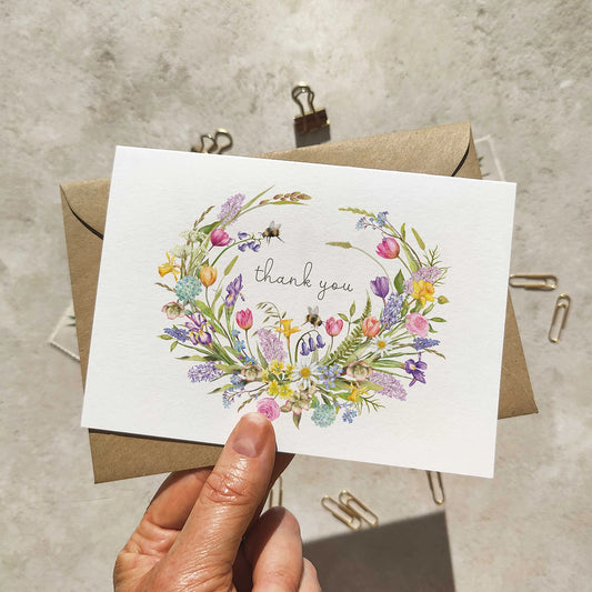 Hand holding a card with a floral wreath and 'thank you' text, placed on a textured surface.