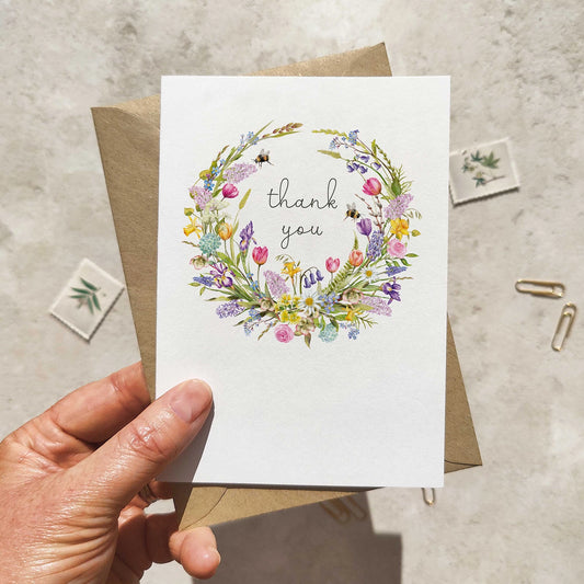 Hand holding a card with a spring floral wreath and 'thank you' text on a textured surface.