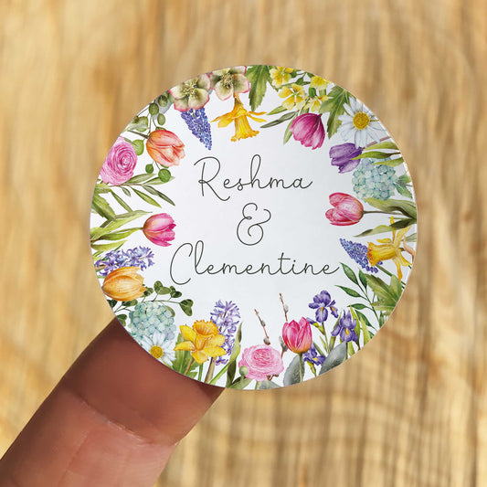 Round sticker with floral design and text 'Reshma & Clementine' held over a wooden surface