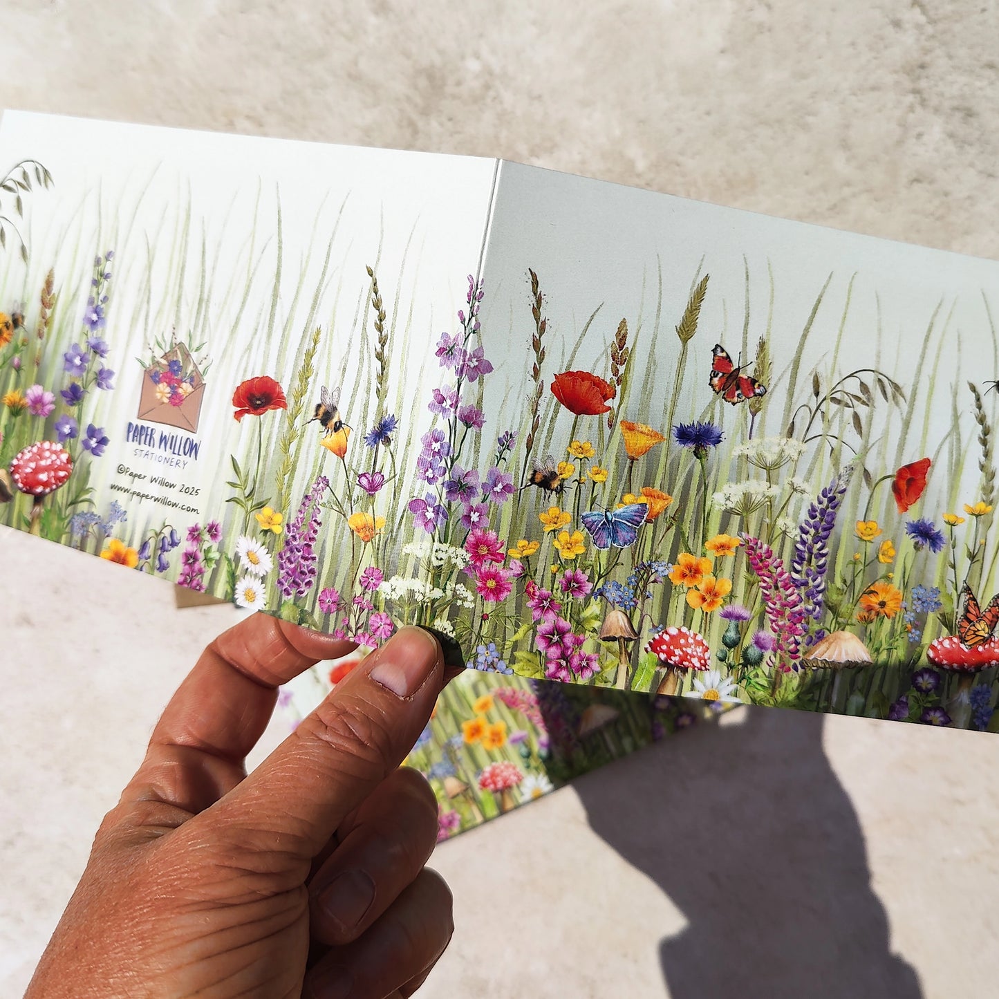 Hand holding a card with a colorful illustration of flowers and butterflies on a textured surface.