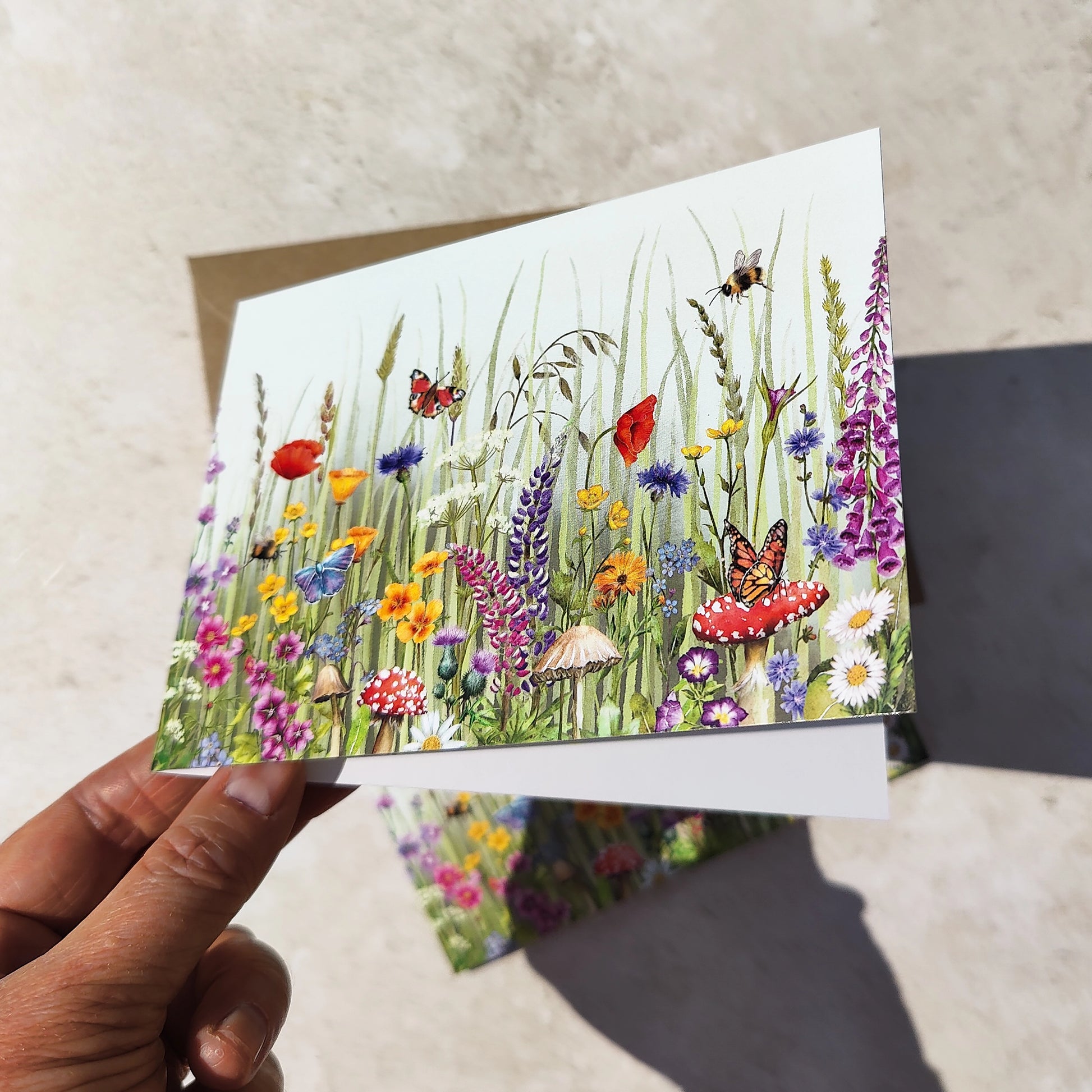 Hand holding a card with a colorful illustration of flowers and butterflies on a textured surface.