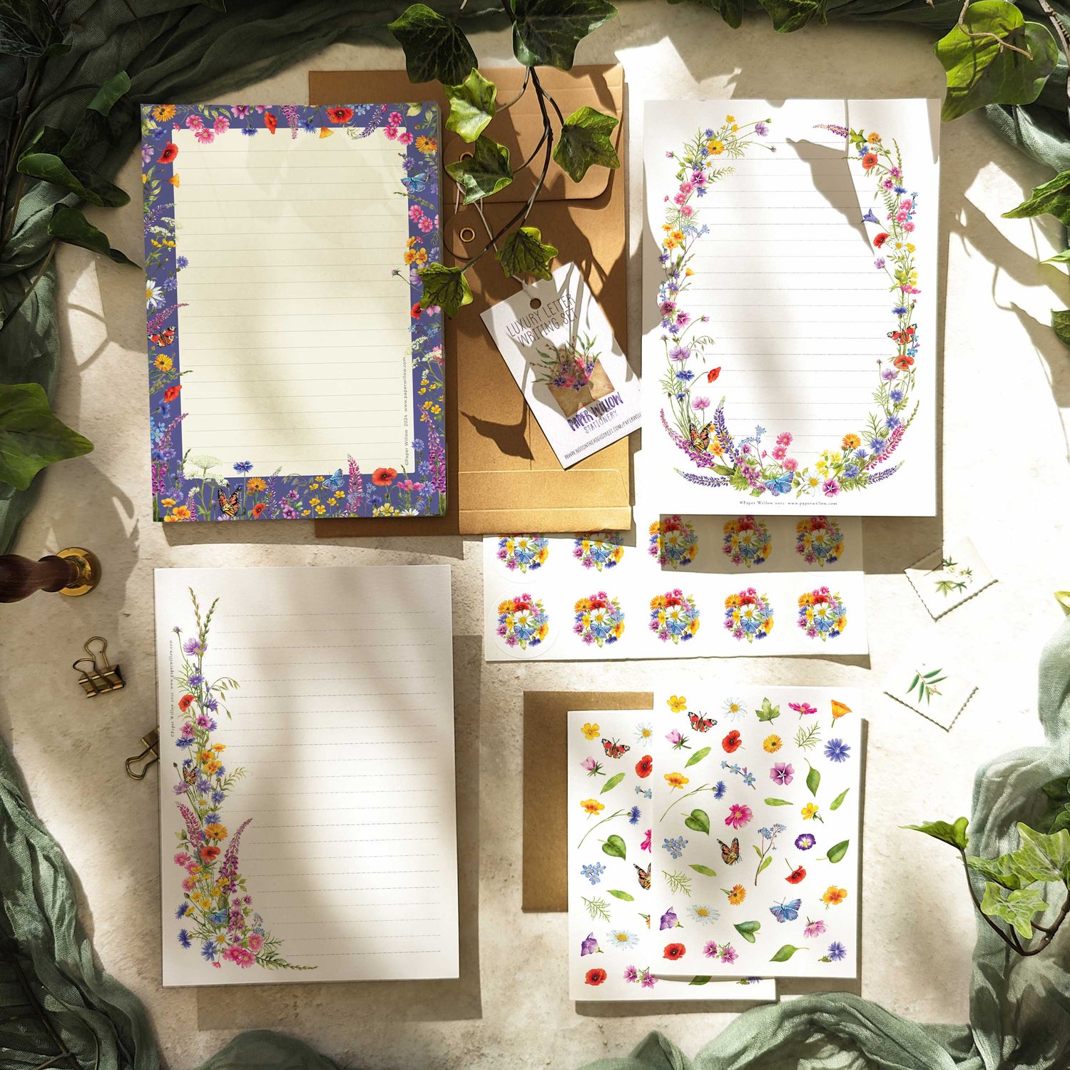 A collection of stationery items with floral designs, including writing paper, notepad, notecards, and envelopes displayed on a table with a green leafy background.