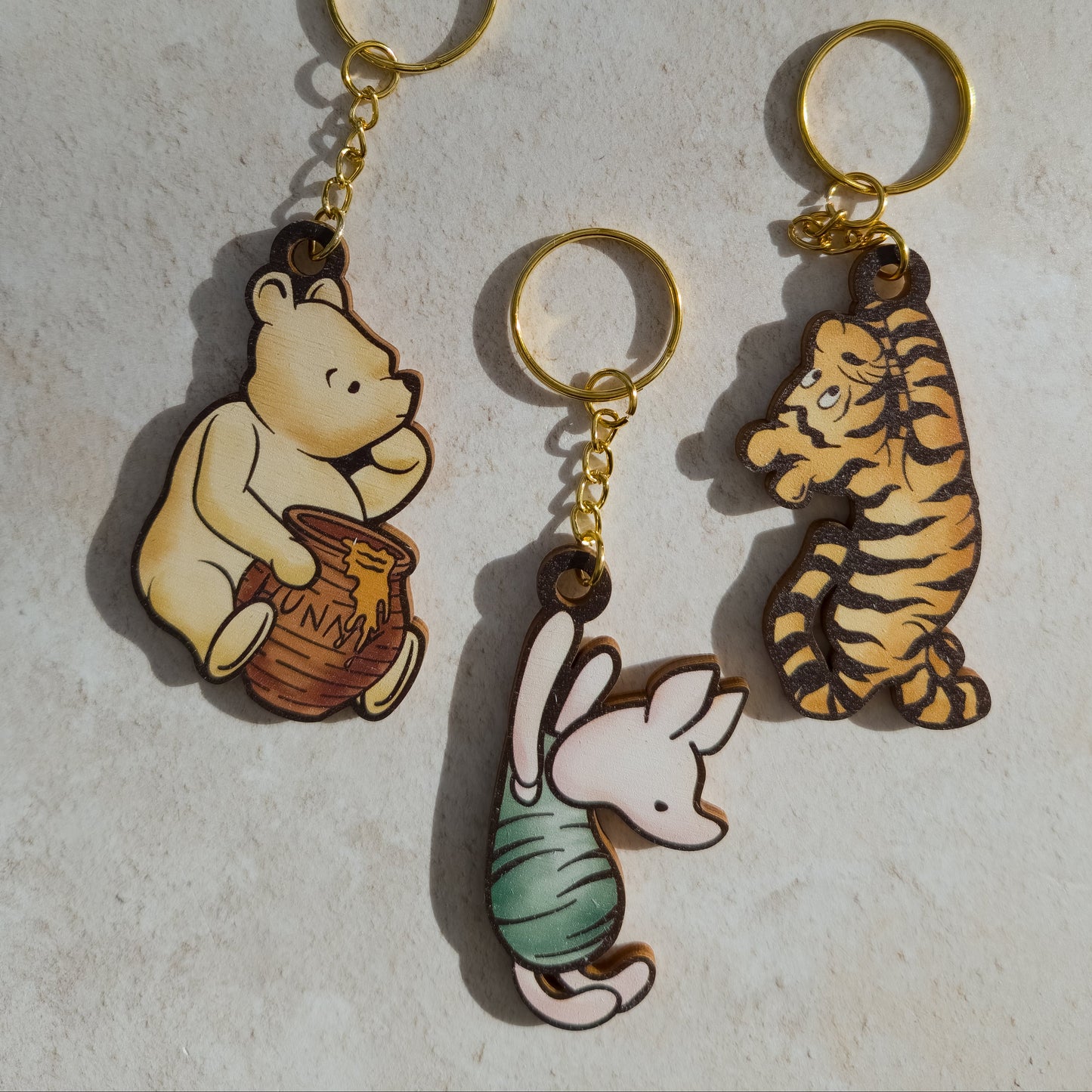 Three character keychains with gold-tone rings and chains, featuring Winnie the Pooh holding a honey pot, Tigger, and Piglet, displayed on a light textured surface.