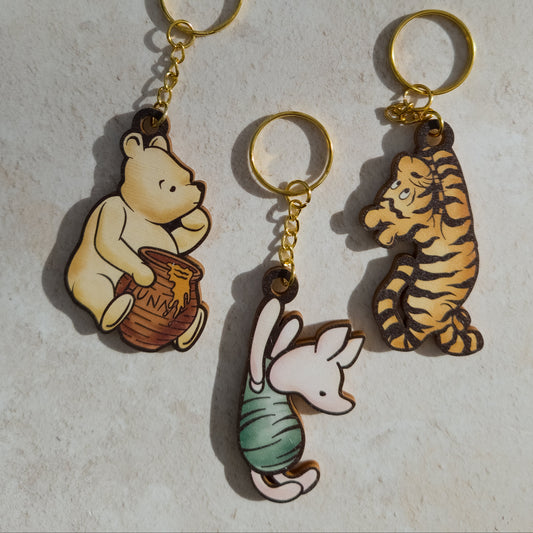 Three character keychains with gold-tone rings and chains, featuring Winnie the Pooh holding a honey pot, Tigger, and Piglet, displayed on a light textured surface.